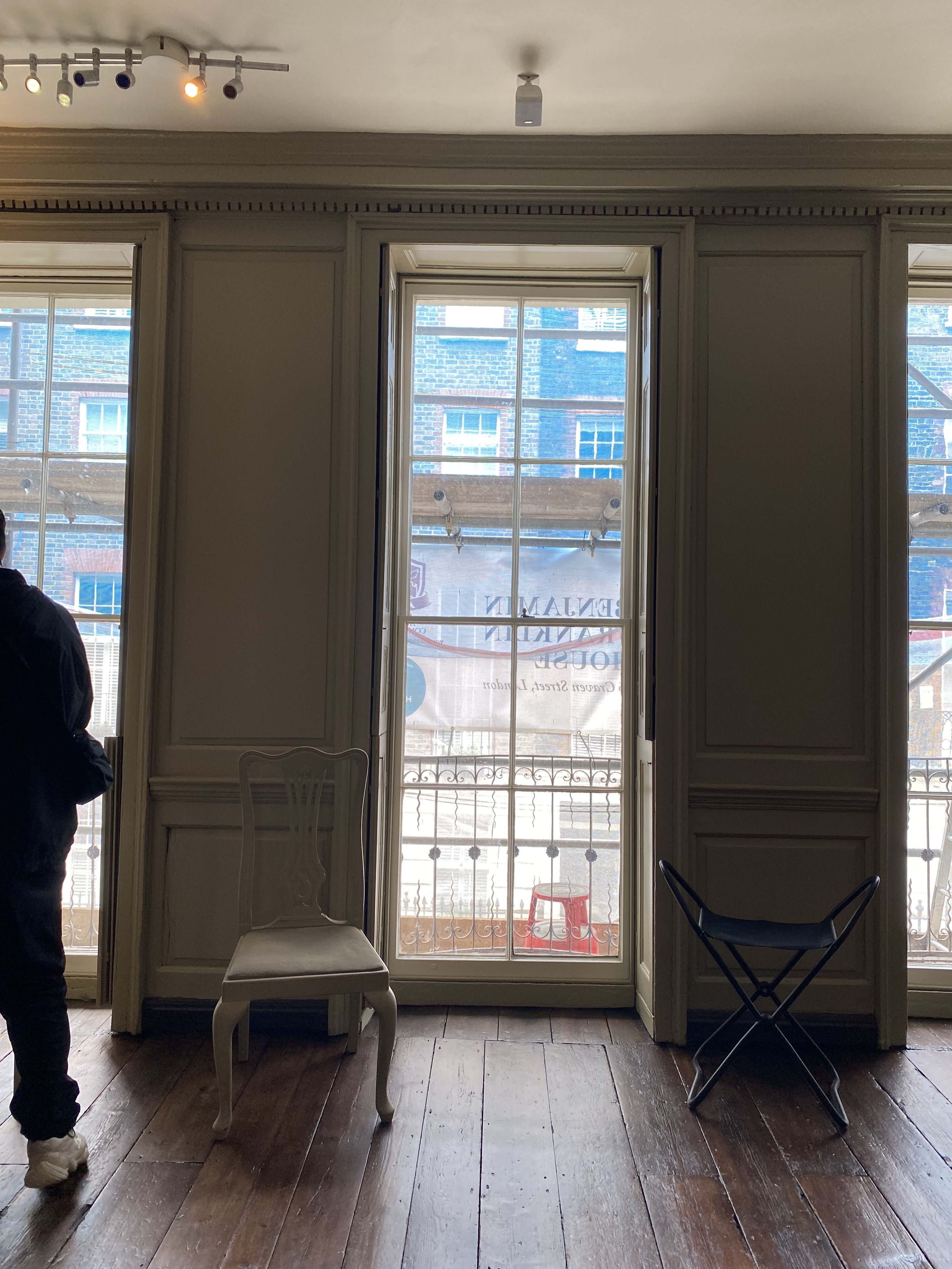 an indoor shot shows a room with a wooden floor and three windows overlooking an adjacent building. a white chair sits near the left window, while a black stool is positioned near the right one. a person is partially visible on the left side of the image.