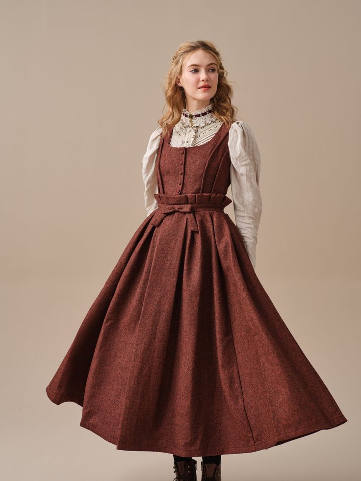 a woman in a long, flowing brown dress is standing in a studio. she is wearing a white lace collar and her hair is styled in loose curls. the dress has a fitted bodice, a full skirt, and a bow at the waist. the background is a neutral beige color. the lighting is soft and diffused, creating a romantic and elegant atmosphere.