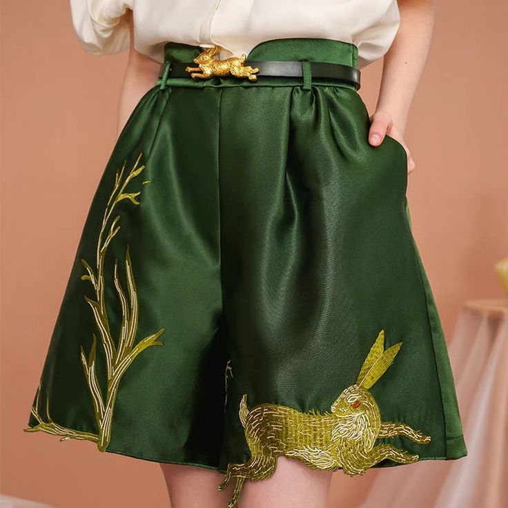 loseup view of a woman wearing a pair of dark green satin shorts. the shorts are highwaisted and have a pleated design.  they feature elaborate gold embroidery depicting rabbits and leafy branches. the woman is also wearing a creamcolored blouse and a black belt with a gold rabbitshaped buckle. the overall style is elegant and sophisticated. the background is a muted peach color with a blurred section suggesting a table or other surface.