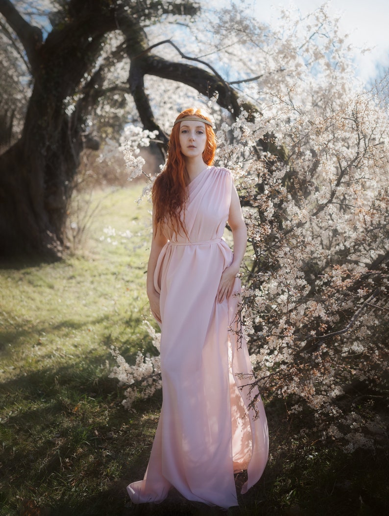 a stunning redhead woman in a flowing pink dress stands amidst a blossoming cherry tree. the image has a dreamy and ethereal quality, with soft lighting and a focus on the woman's beauty and the surrounding natural setting. the overall mood is peaceful and romantic, evoking a sense of fantasy or magic.