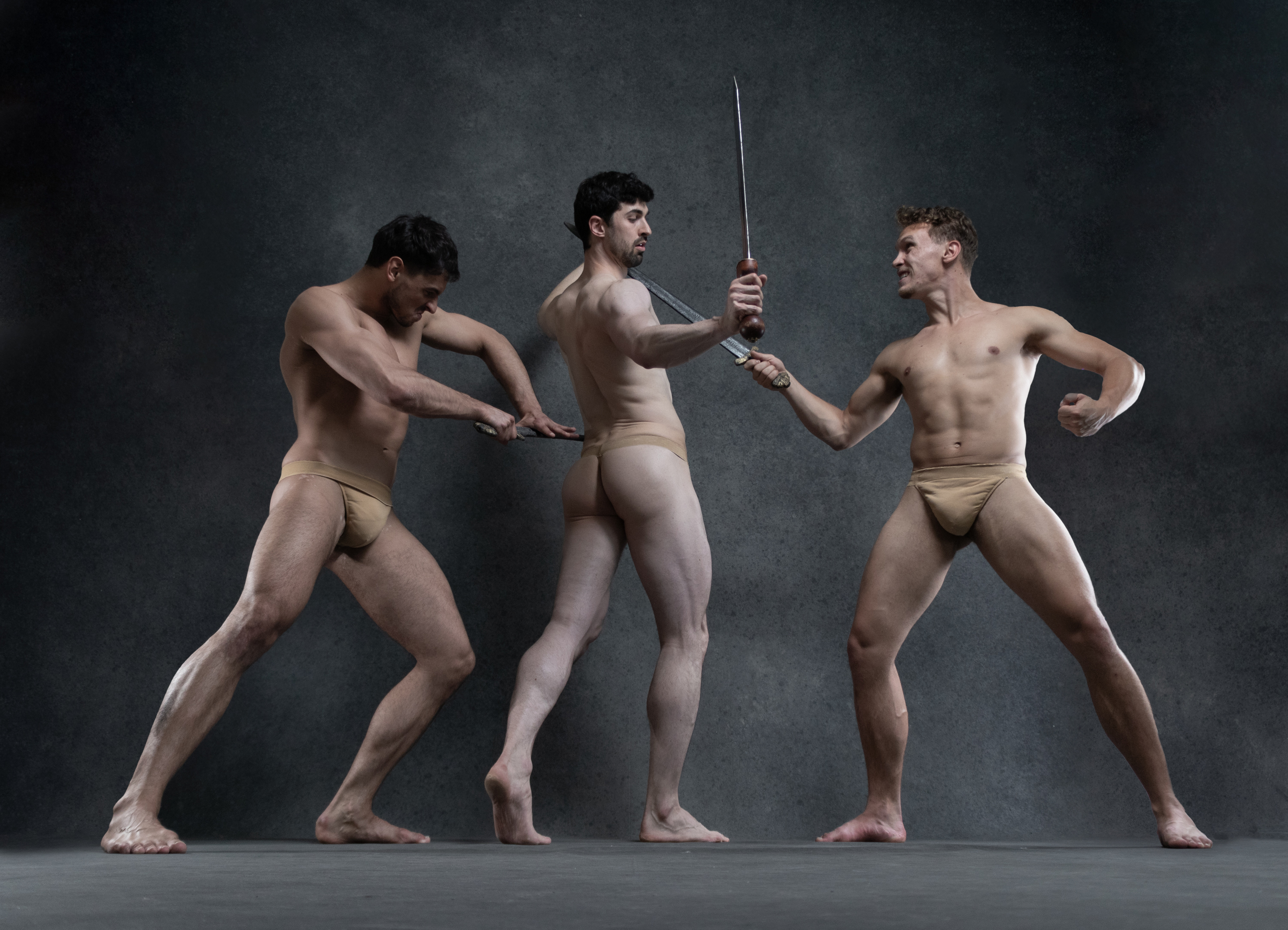 three muscular men in tan thongs are posed in dramatic fighting stances against a dark grey background. the central figure holds a short sword in his upraised right hand. the man to his right appears to be punching with his right fist while the man on the left has his body turned away from the camera and appears to be holding a short sword behind his back.