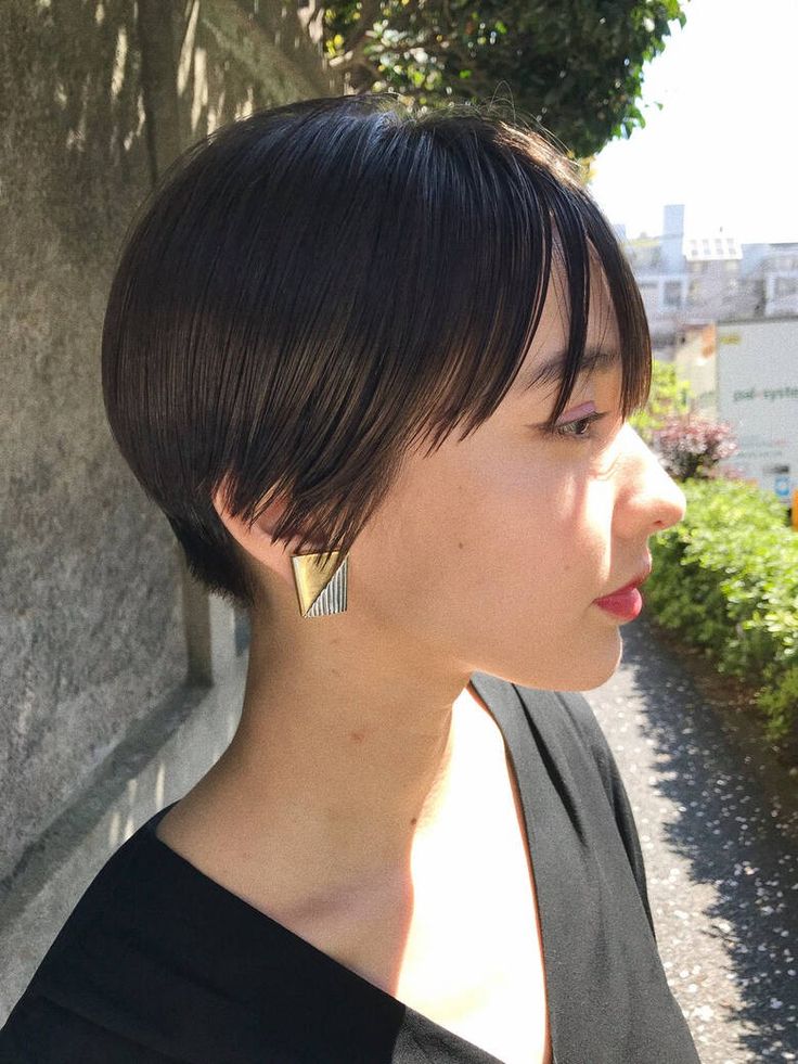 a young woman with short, black hair, wearing a gold and silver earring, is photographed in profile. the woman is looking away from the camera. she is standing on the sidewalk in an urban setting.  the woman's hair is cut in a sharp, angular bob that is slightly longer in the front than the back.  the image is welllit and the woman's face is clearly visible. the image is wellcomposed and focused.