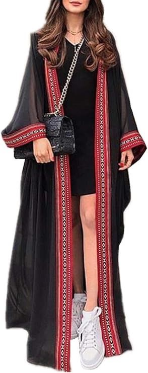 a woman is wearing a black dress with a long, flowing black abaya that has a red and white trim along the edges. she is holding a black handbag with a chain strap and wearing white sneakers. the outfit has a stylish and modern look.