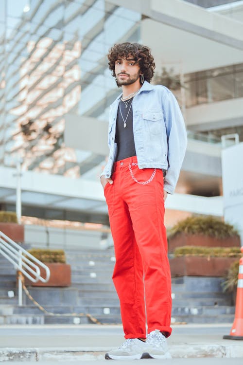 a full shot of a man standing outdoors wearing a denim jacket, trousers, and sneakers. the man has curly hair and a beard, and is wearing a necklace and earrings. the background is a building and an urban scene.