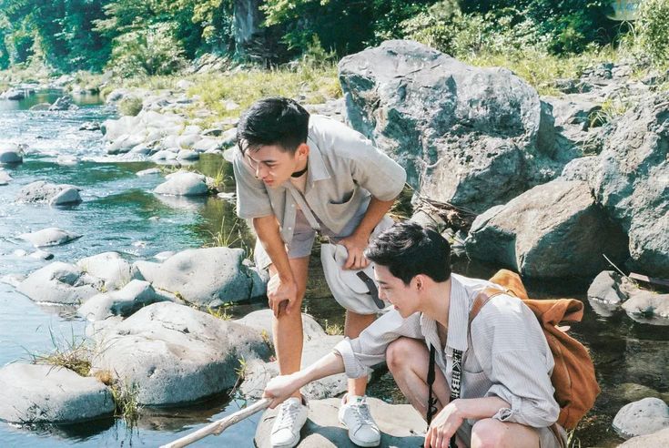 two young men are exploring a rocky riverbed on a sunny day. they are both crouching down, looking at something in the water. one of them is holding a stick. the riverbed is surrounded by lush green forest. the image evokes a sense of adventure, friendship, and connection with nature.