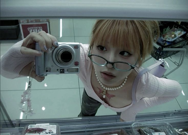 the image is a closeup selfie taken by a young asian woman in a supermarket.  she's wearing a light pink sweater, glasses, and a pearl necklace. a dragonfly tattoo is visible on her chest.  she's taking the picture with a silver compact digital camera, and her reflection is seen in a glass surface (possibly a refrigerated display case). the overall aesthetic is reminiscent of early 2000s fashion trends. the background shows shopping carts and supermarket shelves. the image has a somewhat washedout, cooltoned color palette.