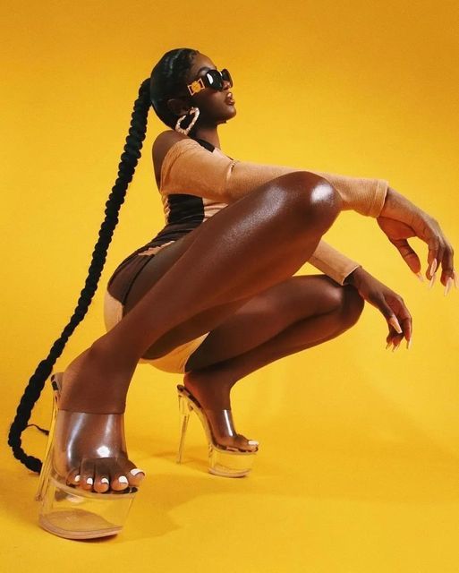 a black woman wearing clear platform high heels with her legs crossed,  posing on a yellow background. she is wearing sunglasses, and her hair is braided.