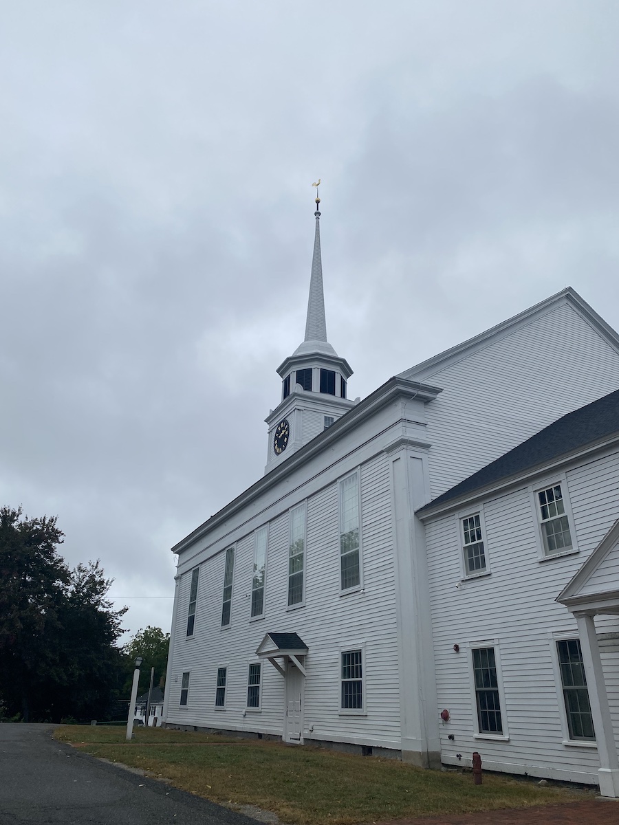 a lowangle shot of a white church on an overcast day. the church has a spire, a clock, and is surrounded by green grass and trees.