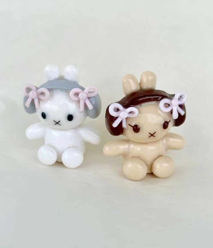 two miniature bunny dolls sit sidebyside. the doll on the left is white with gray ears and the doll on the right is tan with brown ears. both have pink bows on their ears and a stitched "x" for a mouth. they are sitting on a lightcolored surface.