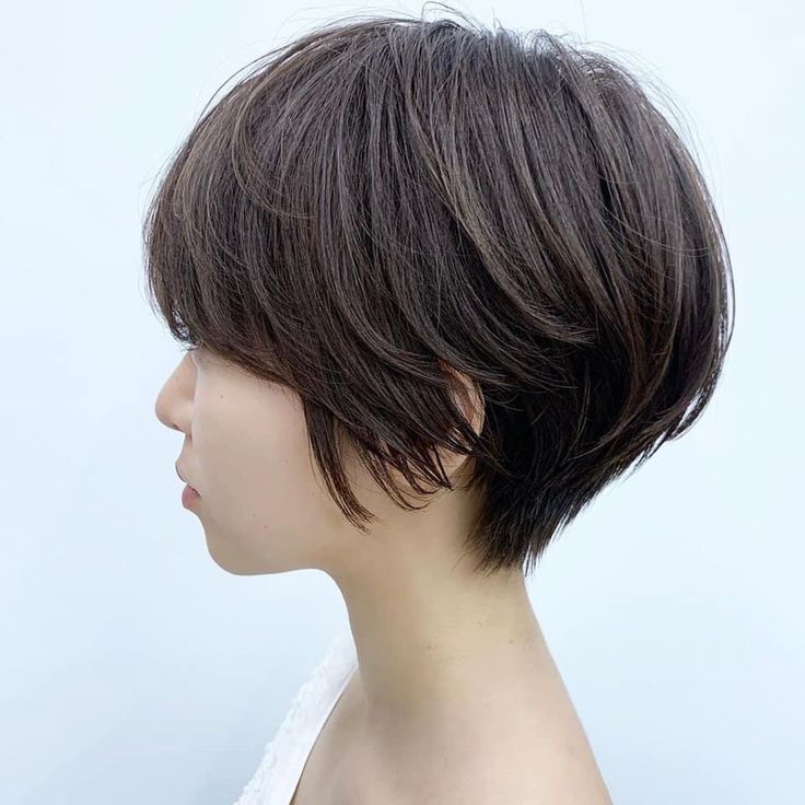 a young woman with short brunette hair, styled in a layered bob, is shown from the side. her hair is textured and slightly messy, and she is looking off to the side. she is wearing a white top.
