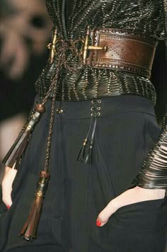 a woman is wearing a black leather dress with a brown leather belt adorned with metallic details. the belt has a tassel on each side. the woman's hands are visible, and one hand is resting in the pocket of the dress. her attire is stylish and chic.