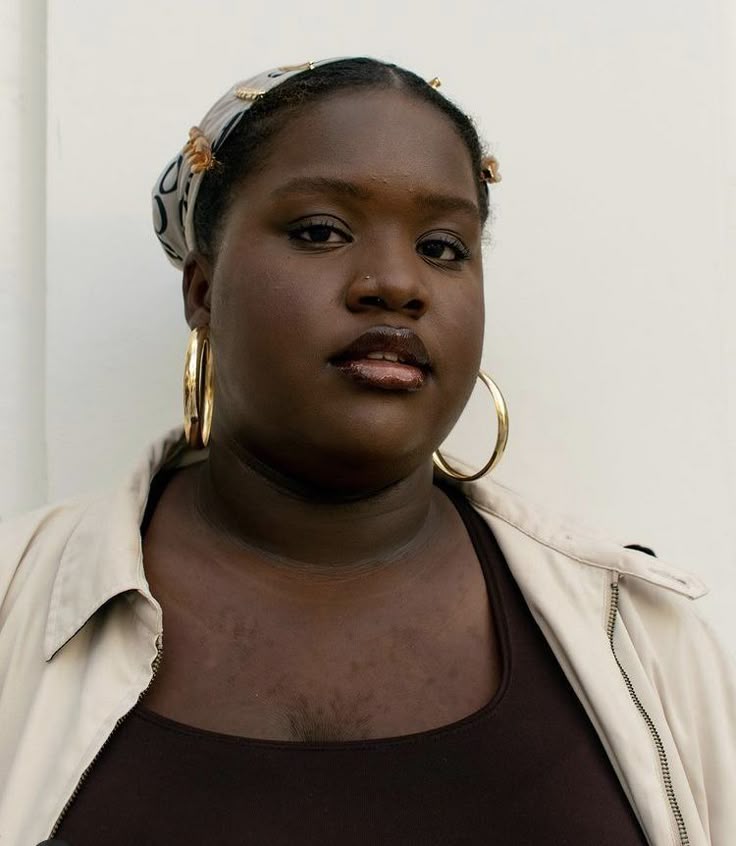 a portrait of a black woman with dark skin, wearing a headscarf, large gold hoop earrings, a brown top, and a beige jacket. she has a nose ring and is looking directly at the camera.