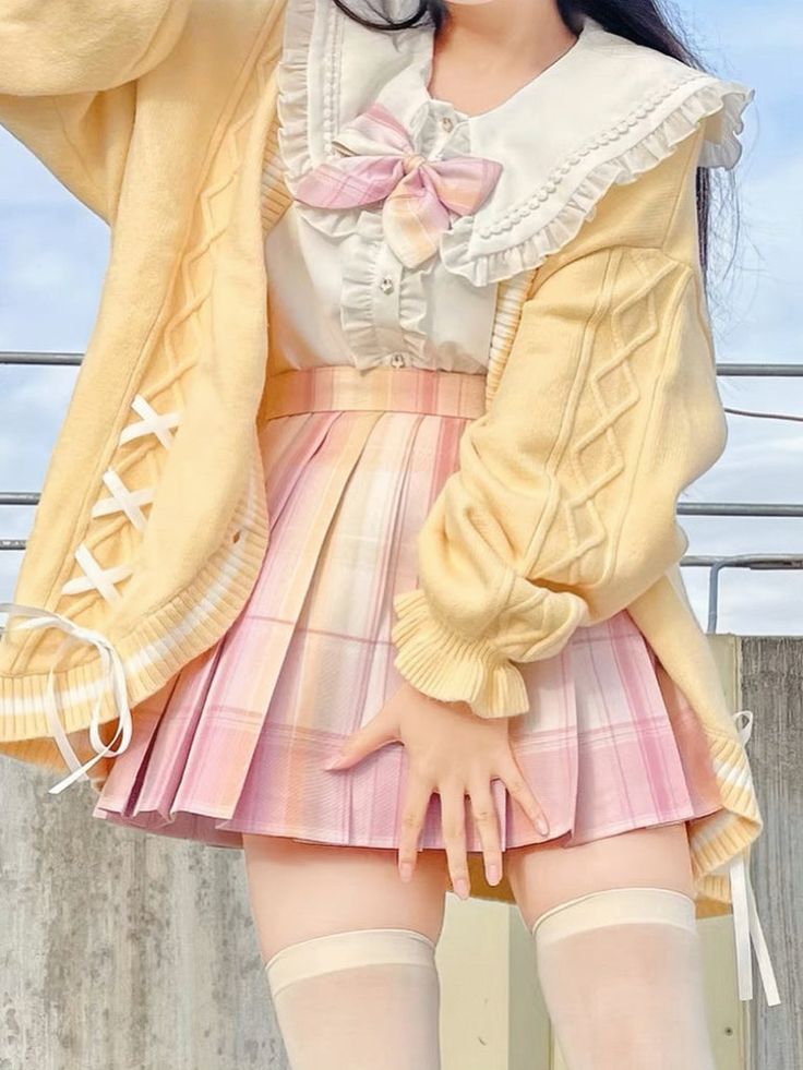 a young woman is standing outside, wearing a pastel yellow cardigan, a white blouse with a lace collar, and a pink and white plaid skirt. she is wearing white thighhigh socks. her outfit is in the lolita style, a japanese fashion subculture known for its dolllike aesthetic. the image is taken from an eyelevel perspective and is welllit by natural light.