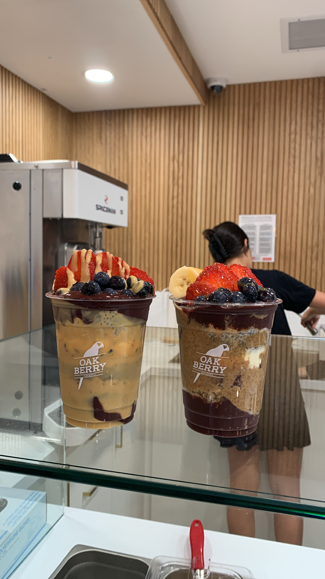 two oak berry acai bowls are on a counter, topped with fruit and granola. a woman in a black skirt and white tshirt is working behind the counter. the restaurant interior has light wood paneling.