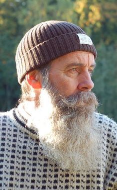 a closeup shot of a man wearing a brown knit hat and a patterned knit sweater. he has a long, mostly white beard and mustache. the background is blurred greenery, suggesting an outdoor setting.