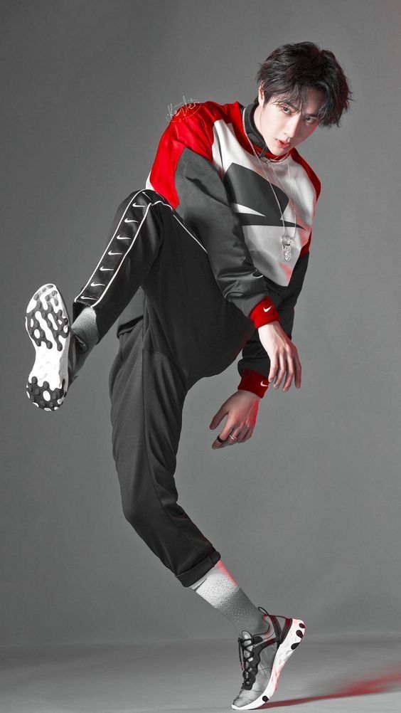 a young man in stylish red and black nike sportswear strikes a dynamic dance pose against a minimalist gray backdrop. the studio lighting highlights his form and the fluidity of his movement. he wears black track pants with white accents, a red and white colorblocked sweatshirt, and black and white nike sneakers. his expression is focused and intense, showcasing the energy and confidence of his pose.