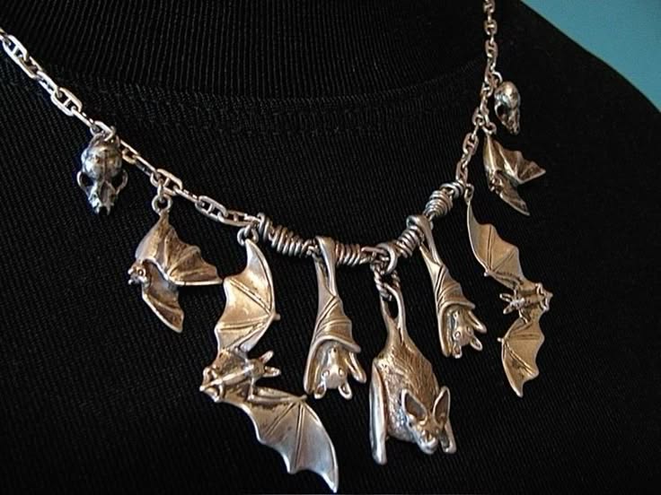 a closeup of a silver necklace with multiple bat pendants. the necklace is on a black shirt. the bat pendants are all different sizes and shapes, and they are all hanging from a silver chain. the necklace is intricate and detailed, and it appears to be handmade. the necklace is a spooky and stylish accessory that would be perfect for a gothic or halloween look.
