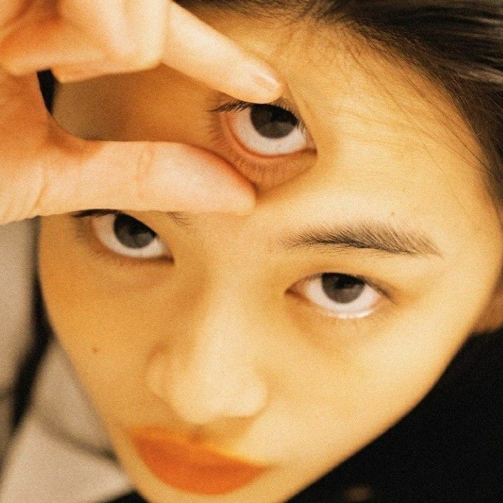 a closeup portrait of a young woman with her hand covering one eye. the woman is looking directly at the camera. her face is illuminated by a warm, yellow light. her eyebrows are dark and defined. her nose is straight and slightly upturned. her lips are painted in a bright orange lipstick. the woman's hand is covering her left eye. her fingers are spread apart, and her thumb is resting on her forehead. the woman's hand is partially visible in the frame, and her skin is smooth and flawless. the background is blurred and out of focus. the overall impression of the portrait is one of beauty and intrigue.
