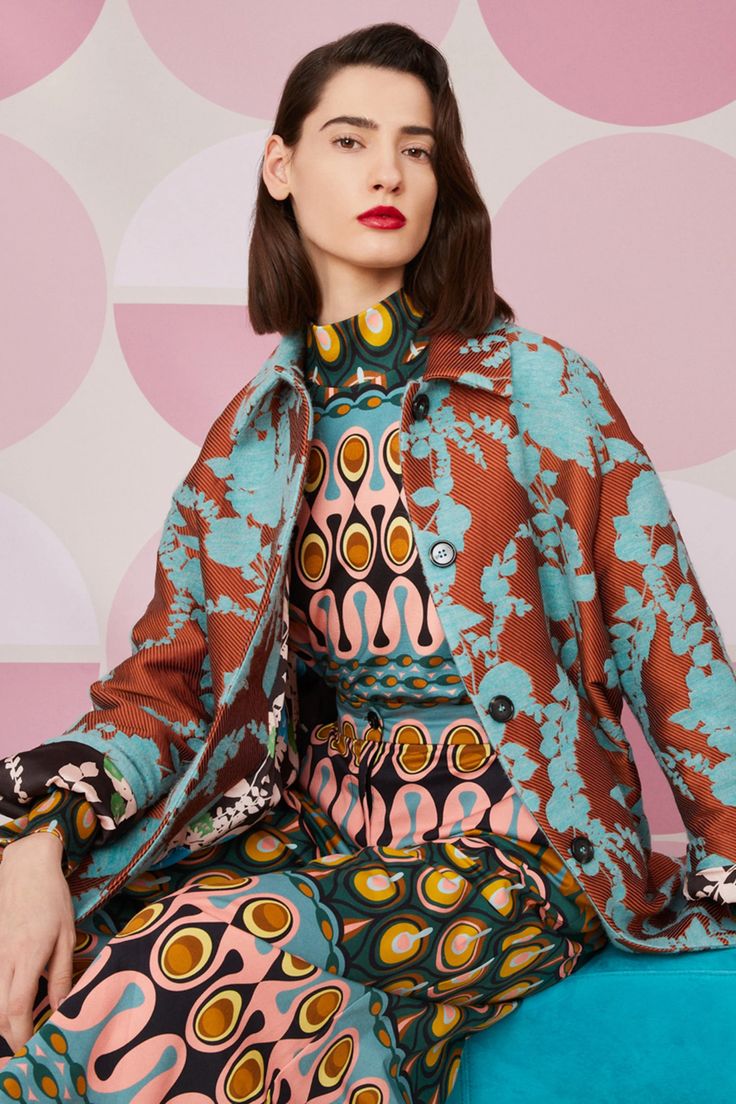 a fashion model with shoulderlength brown hair is posing while wearing a striking outfit. the ensemble consists of a teal and rustcolored floral printed jacket worn over a matching jumpsuit featuring a vibrant, retroinspired psychedelic print combining teal, pink, orange, and black.  the model's red lipstick and confident pose complement the bold design of the clothing. the background features large pastel pink circles on a white backdrop. the overall image evokes a sense of high fashion and vintage style.