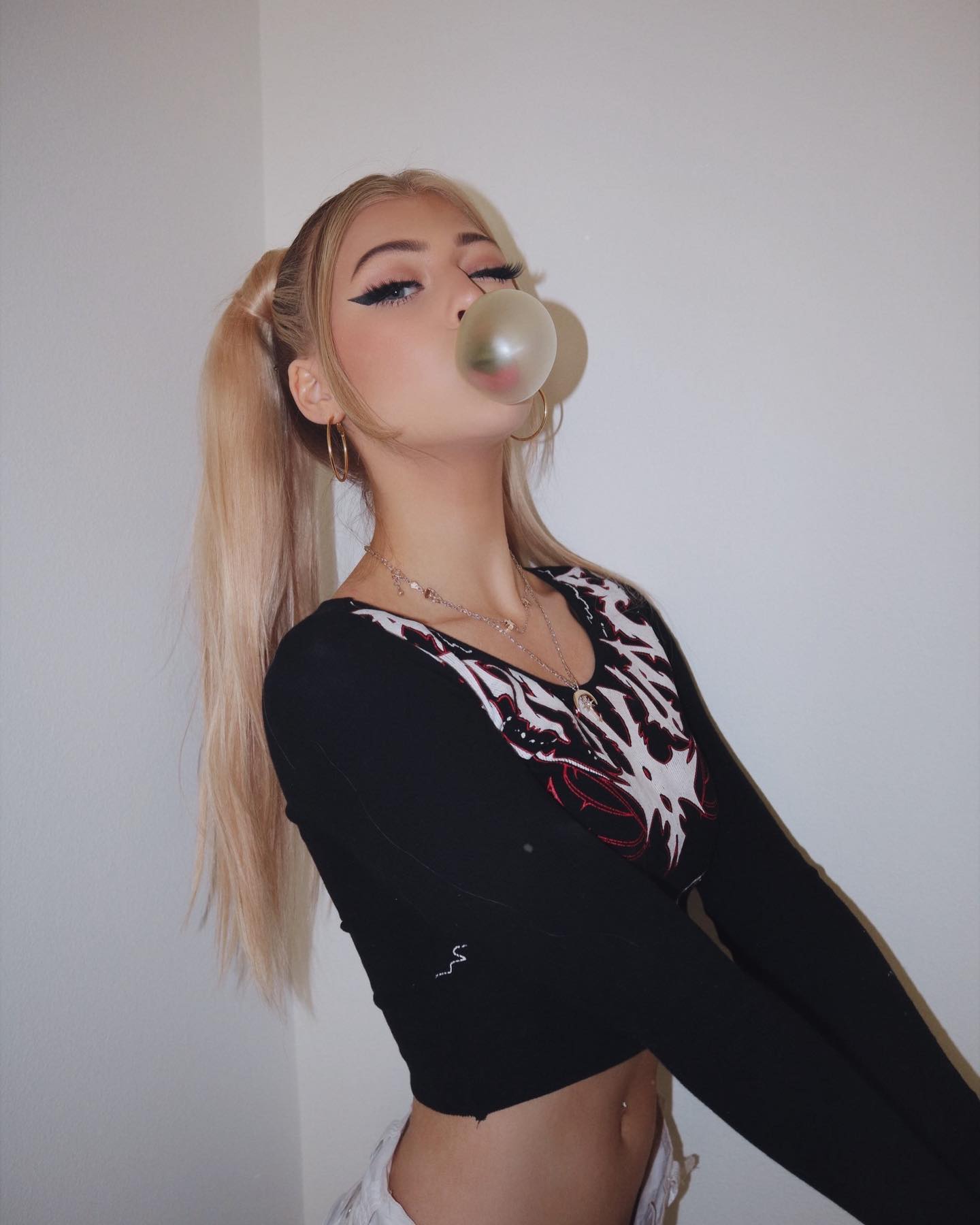 a young woman with blonde hair pulled into pigtails is posing indoors against a white background. she is blowing a bubble with gum. she is wearing a black longsleeve crop top with a unique graphic design on the chest, hoop earrings, and multiple necklaces. her makeup includes eyeliner and she has full lips. a navel piercing is visible. she appears confident and is dressed in a stylish fashion.