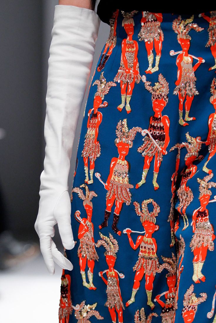 a close up shot of a model on the runway showcasing a unique and eyecatching dress. she is wearing white gloves, and the dress features a vibrant blue fabric with colorful, embroidered figures. the detail and craftsmanship of the print are evident, creating a visually striking and sophisticated look.