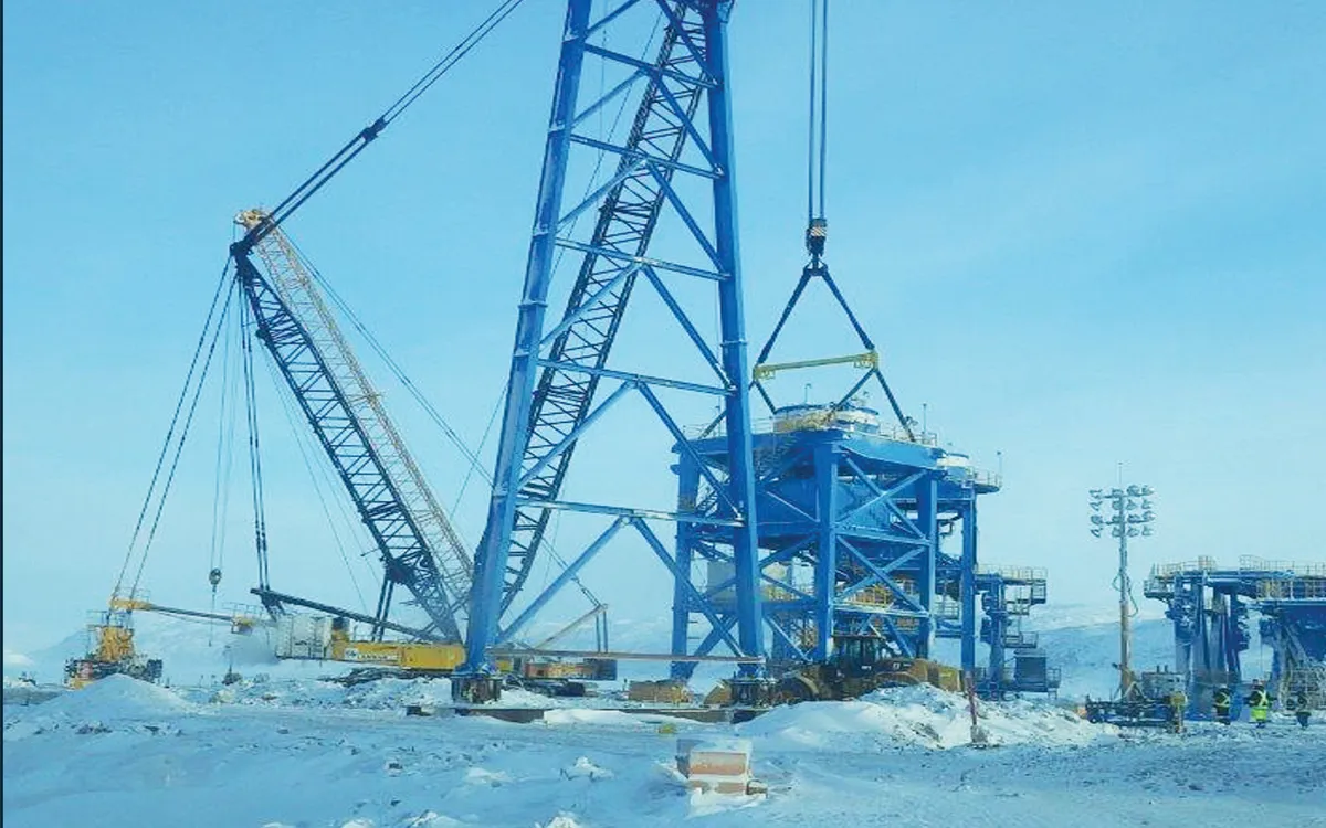 a large blue crane is lifting a portion of a construction project.  the crane is set on a snowy ground with a blue sky in the background.  another crane is visible to the left and there is a structure under construction to the right. the equipment is likely being used to assemble a large industrial structure.