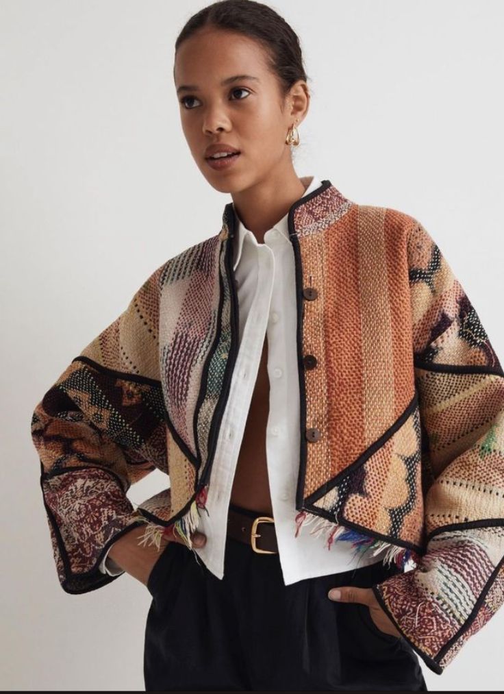 a woman is wearing a colorful patterned jacket with a white buttondown shirt underneath and black pants. she has her hands in her pockets and is looking at the camera. the jacket is a patchwork of different fabrics and patterns and has a brown belt at the waist.