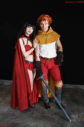 a full shot of a couple in cosplay, portraying characters from a fantasy game. the woman is wearing a red cloak and a red and gold dress, while the man is in a yellow tunic and red pants. they're holding a large sword together. the backdrop is dark, focusing on the couple.