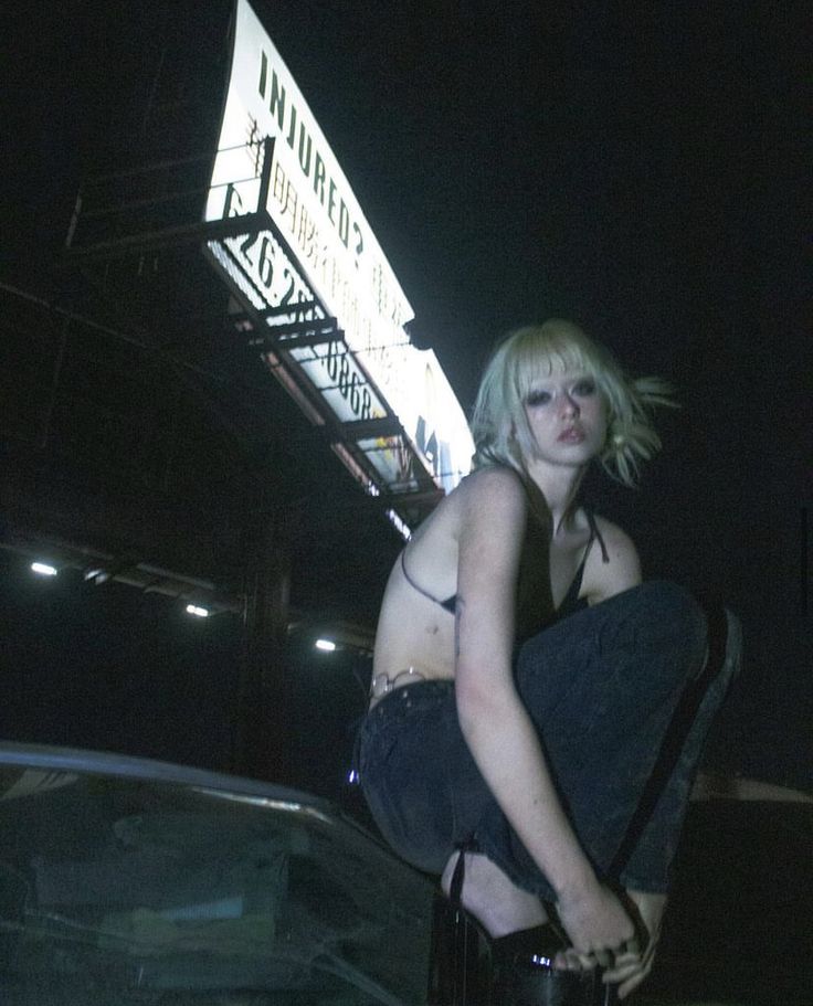 the image is a moody, nighttime portrait of a young blonde woman with short hair. she's dressed in dark, edgy clothing – jeans, a dark top, and platform shoes – and is sitting on the hood of a car.  above her is a large billboard with the word "injured?" prominently displayed. the overall atmosphere is dark, urban, and somewhat mysterious, with a grunge or alternative aesthetic. the scene is set at night, and the lighting is low and dramatic, focusing primarily on the woman.  the overall vibe suggests a rebellious or independent spirit.