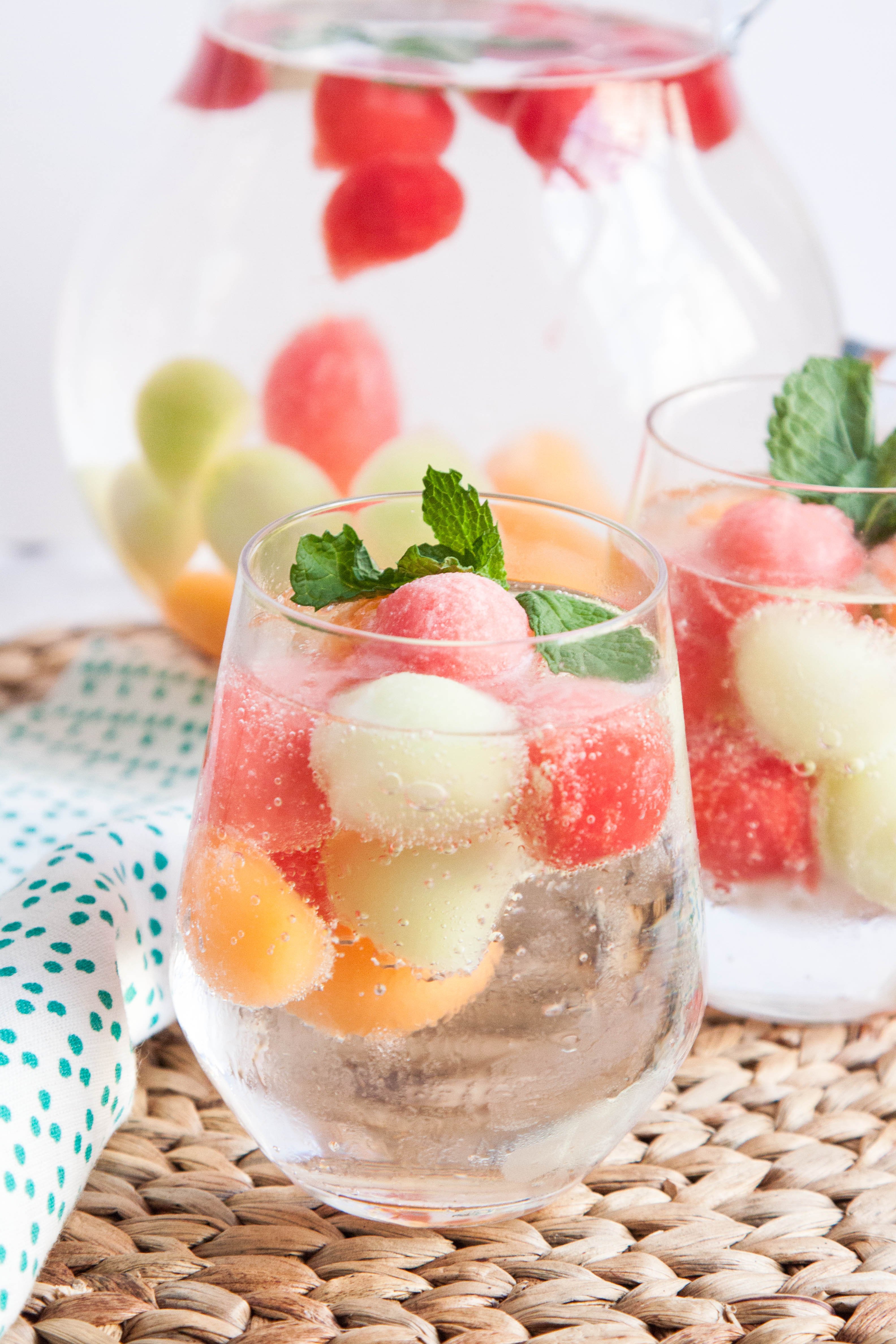 two glasses filled with a refreshing summer beverage. the drink contains frozen watermelon, cantaloupe, and honeydew melon balls, creating a visually appealing and flavorful mix.  fresh mint sprigs garnish the glasses, adding a touch of elegance. the glasses sit on a woven placemat, and a large pitcher filled with more of the same fruit ice is visible in the background. the image evokes a feeling of summer refreshment and relaxation.