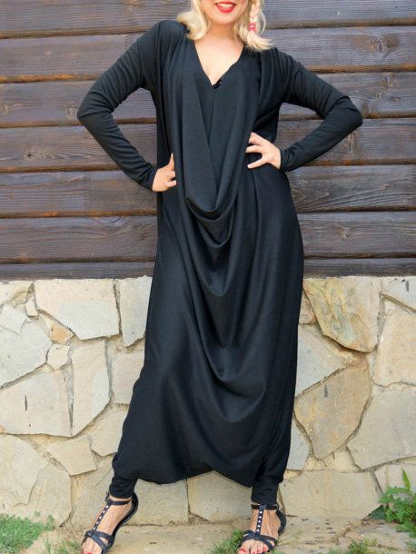 a woman stands in front of a stone and wood wall, wearing a black draped jumpsuit with long sleeves. she has blonde hair and is wearing red lipstick and sandals. she also has jewelry on her left ear.
