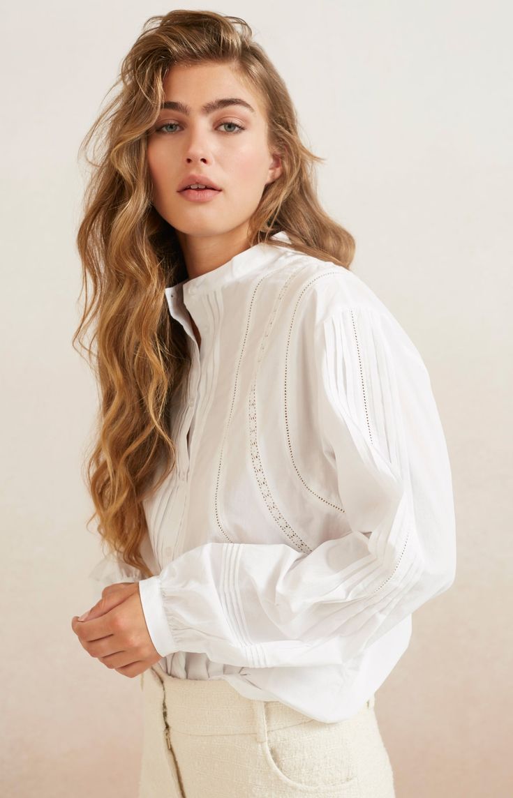 a woman with long blonde wavy hair, wearing a white blouse with long sleeves and a white collar. she is looking at the camera. the image is a portrait taken in a studio. the woman is wearing minimal makeup and the lighting is soft.  the background is white. the woman appears to be in her 20's.