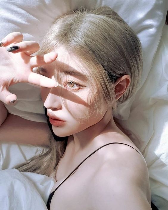 a woman with blonde hair is lying in bed with her eyes partially obscured by her hand. the sunlight is casting shadows across her face and body, highlighting the contours of her features.