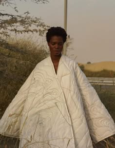 a woman with short dark hair wears a white, quilted, robelike garment in a desert environment. the garment appears to be windblown and flowing. the image is shot in a way that emphasizes the texture of the fabric, and the woman is looking down as if in thought.