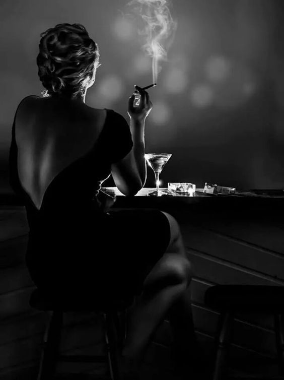 a woman sits at a bar in a dimly lit setting. she holds a lit cigarette in one hand and is dressed in a black sleeveless dress.  her back is turned towards the camera, and she is looking over her shoulder. there is a cocktail glass on the counter in front of her. the image is in black and white, creating a sense of mystery and intrigue.