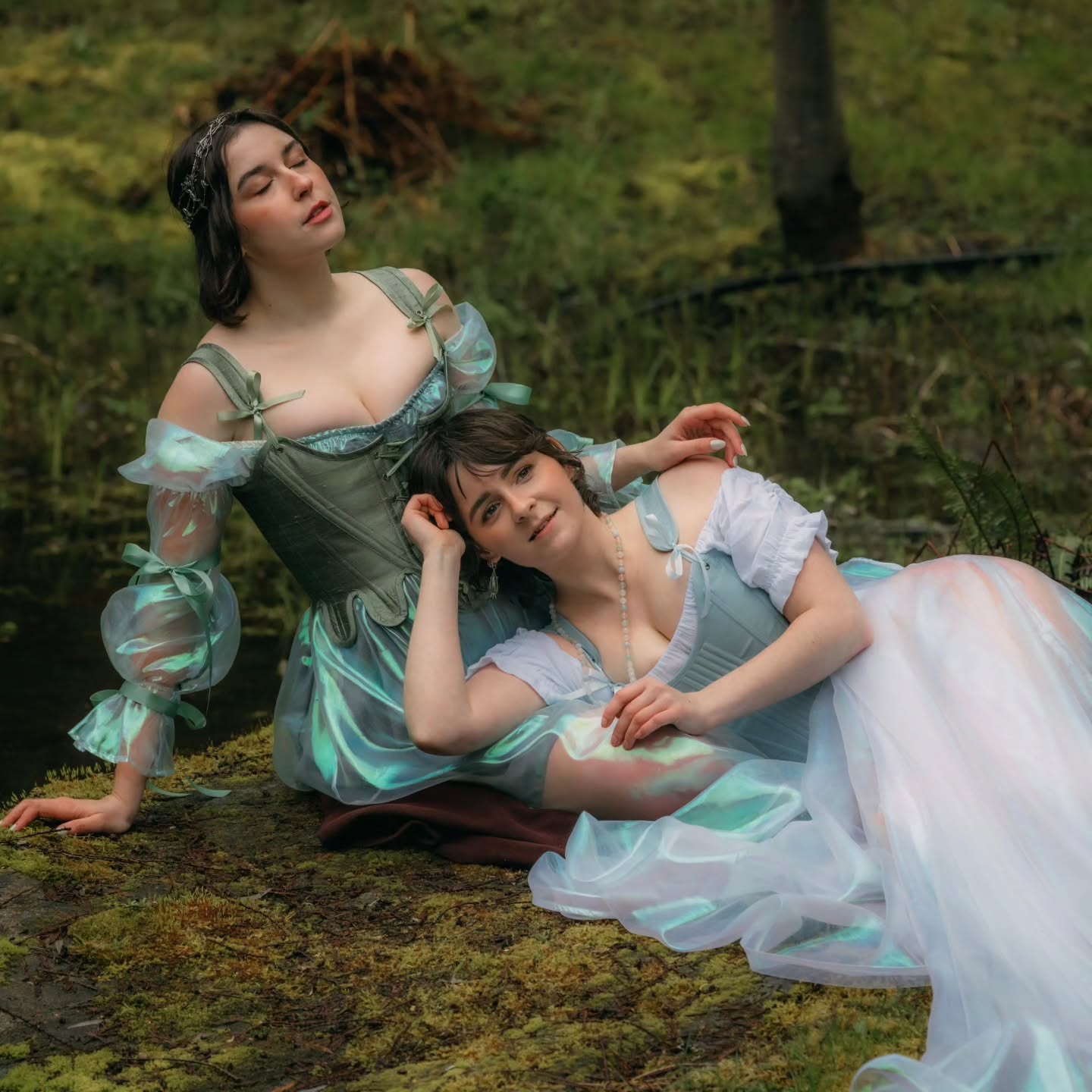 two women in elaborate teal and light blue renaissancestyle dresses are captured in a picturesque outdoor setting.  they are nestled amongst mossy rocks and greenery in what appears to be a forest. one woman sits with her arm around the other, who is lying down, creating a sense of intimacy and companionship. the dresses are detailed and flow beautifully, enhancing the ethereal and romantic mood. the overall scene conveys a feeling of fantasy and whimsy.