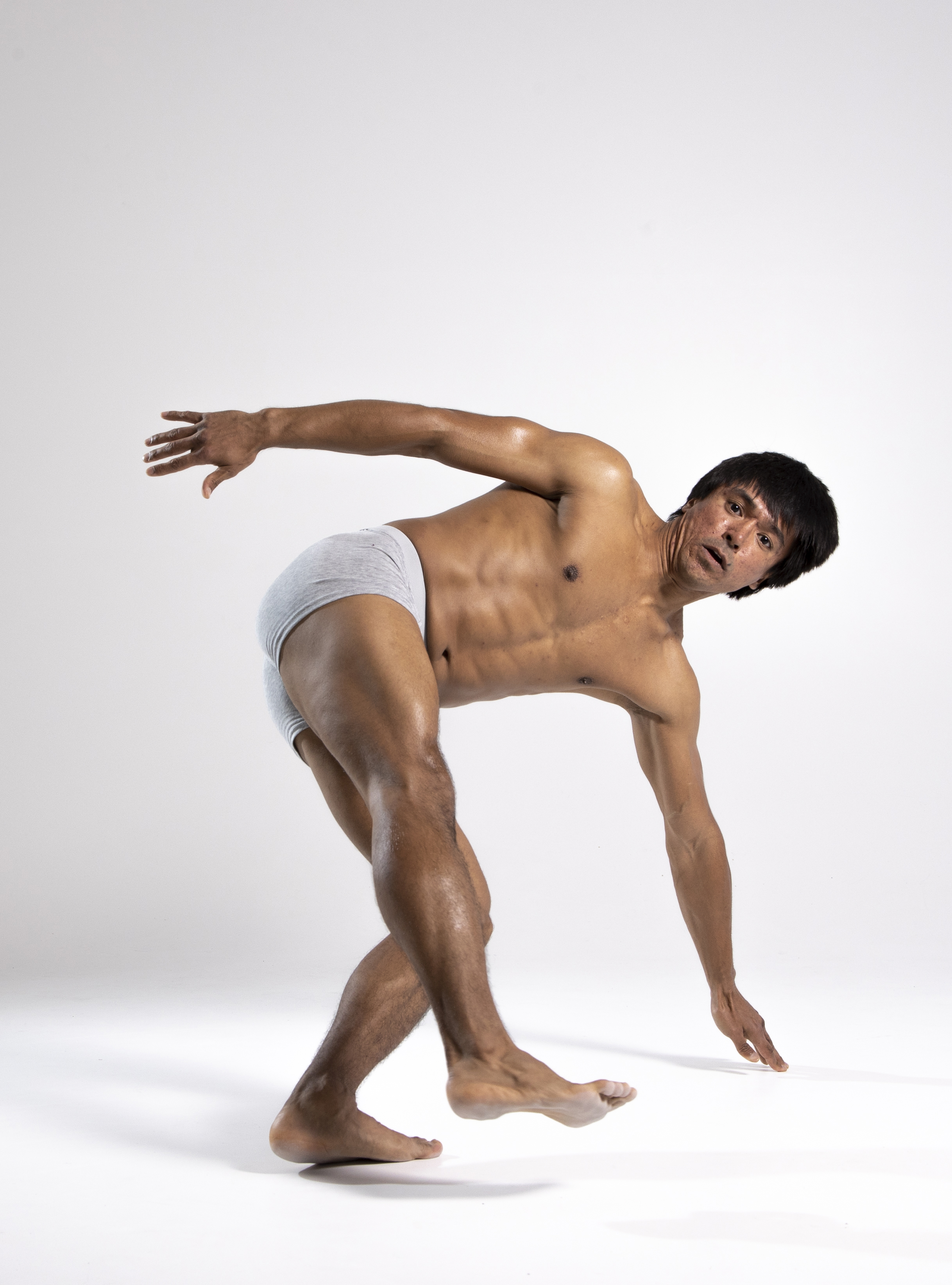 a muscular, shirtless man wearing underwear is posing in a studio. he is barefoot and leaning forward with one hand extended towards the ground. he is looking at the camera with a focused expression, flexing his physique. the background is a plain white studio backdrop.