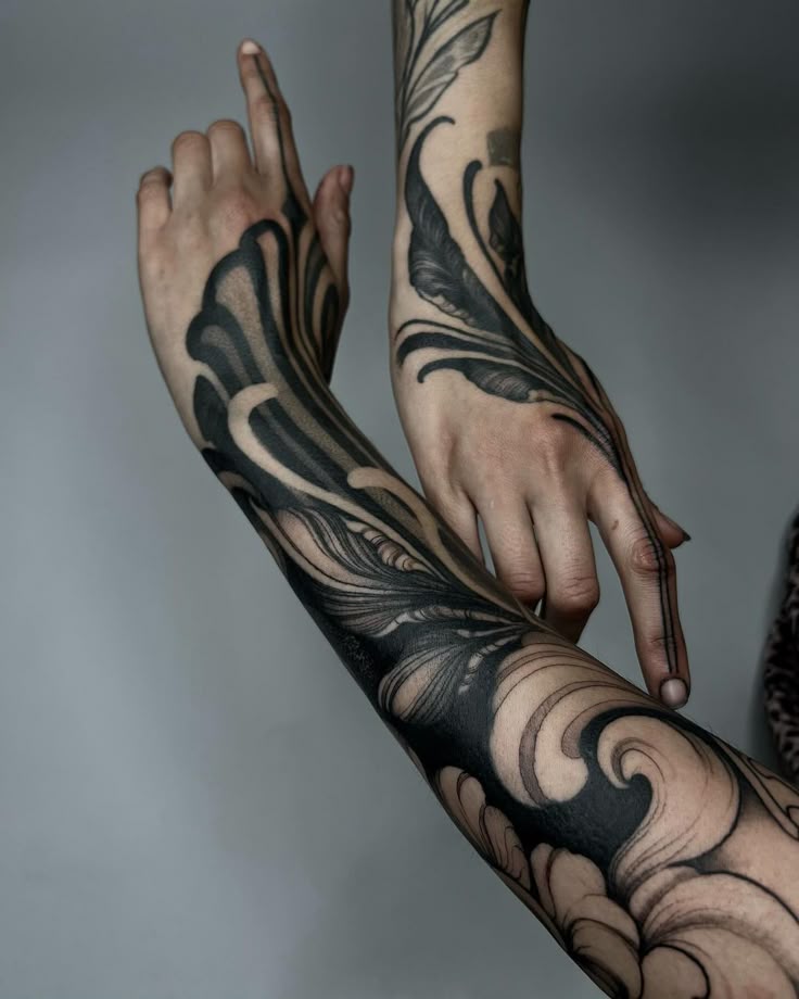 this image showcases two arms adorned with intricate black tattoos. the left arm features a prominent wave tattoo design that extends from the forearm to the hand. the right arm has a delicate leaf tattoo pattern. the tattoos are highly detailed, featuring elegant lines and shading. the person's hands are positioned in a way that highlights the design. the tattoos cover a significant portion of their arms, adding a unique and artistic touch to their appearance. the black ink stands out against the skin, creating a striking contrast.
