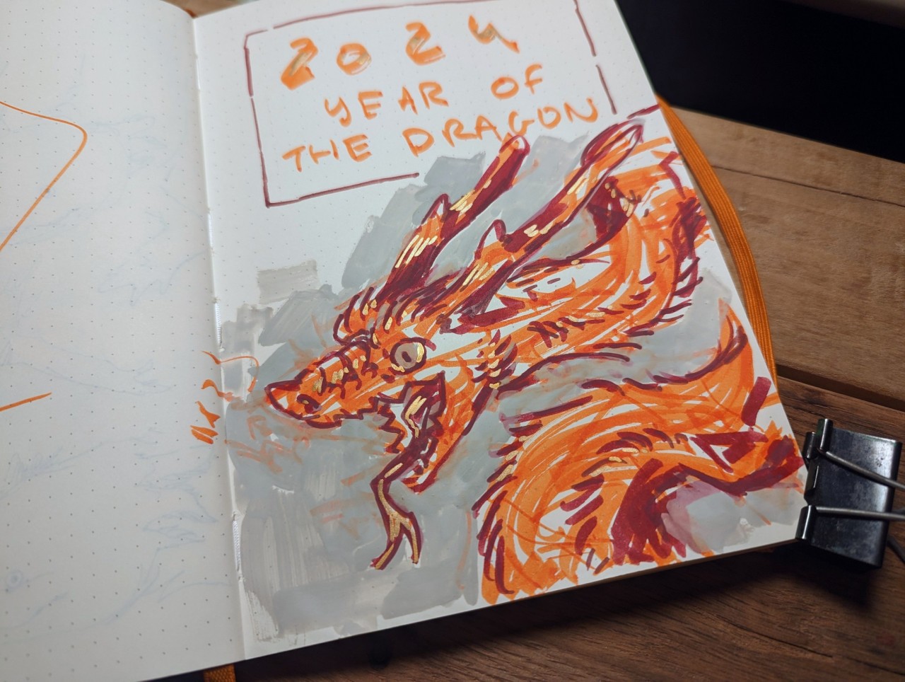 a sketchbook page open to a drawing in orange and grey of a dragon's head and part of its body. the dragon is breathing fire or smoke towards the left, and is facing the left. it's drawn in a slightly cartoonish style. there is text above the dragon that says "2024 year of the dragon".  the image is lit with artificial lighting.  the drawing has been created in a dot grid notebook with an orange elastic closure band. a black binder clip is also visible.