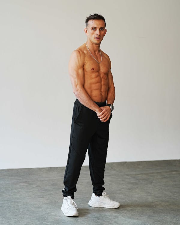 a muscular man is standing shirtless in a studio, wearing black pants and white sneakers. he has a silver chain around his neck and a wristwatch on his left wrist. he is looking at the camera with a neutral expression. the background is a plain gray wall and floor.