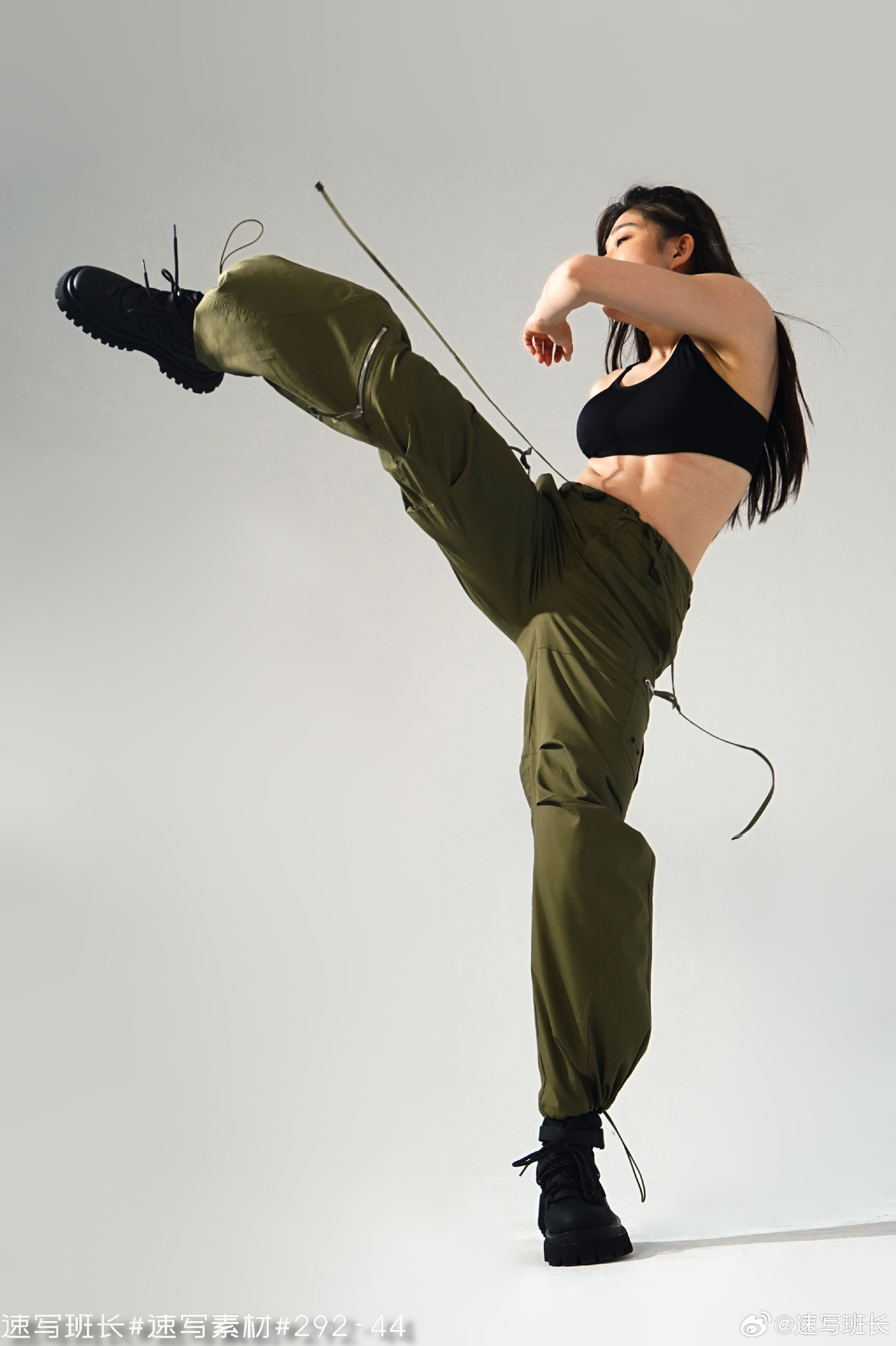 a woman with a toned physique is captured midair executing a powerful high kick.  she wears black athletic top and loosefitting green cargo pants, highlighting her athleticism.  the studio setting with bright artificial lighting creates a strong contrast and emphasizes the dynamic motion. the low angle perspective further accentuates the power and height of her kick.