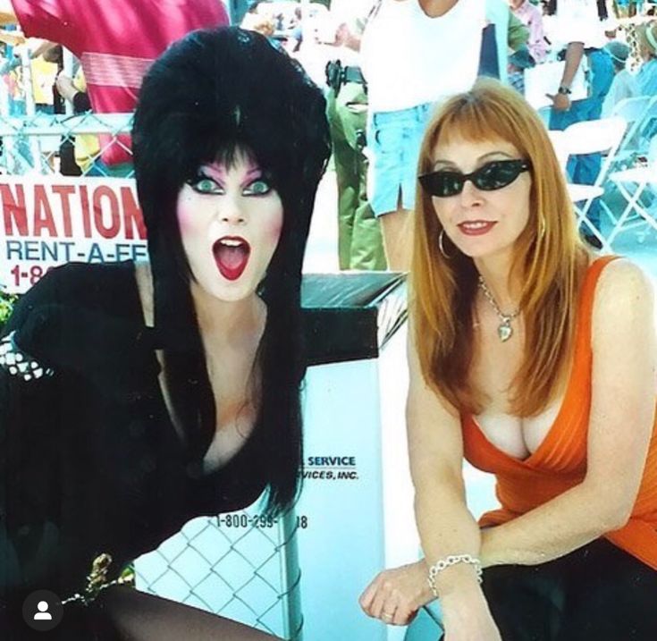 a photo of lisa loring, the actress who played wednesday addams in the addams family, at an event. she is wearing a black dress and smiling, and another woman is standing beside her. the woman beside her is wearing an orange top and has red hair.