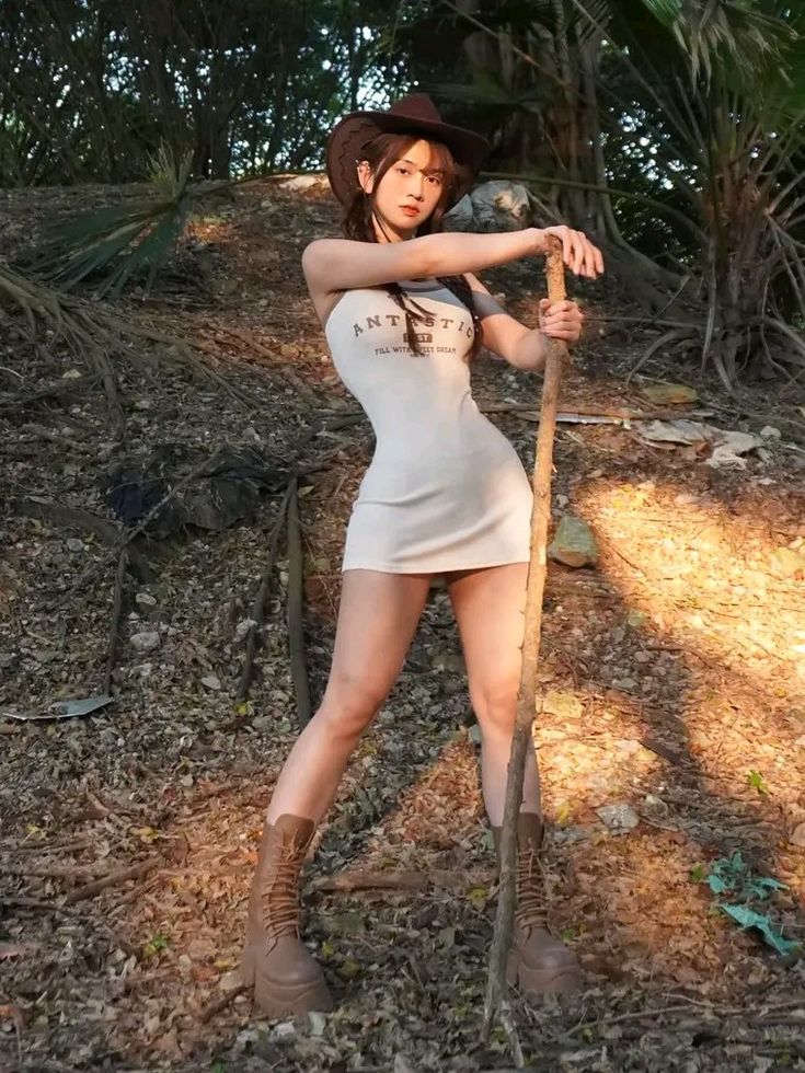 a young woman with long brown hair, wearing a cowboy hat and a short beige dress, stands in a wooded area. she holds a long stick or staff.  she's wearing brown combat boots and her pose is somewhat casual, yet striking. the sun shines on her, illuminating parts of the scene and creating a slightly shadowed forest backdrop. the overall impression is one of casual outdoor fashion.