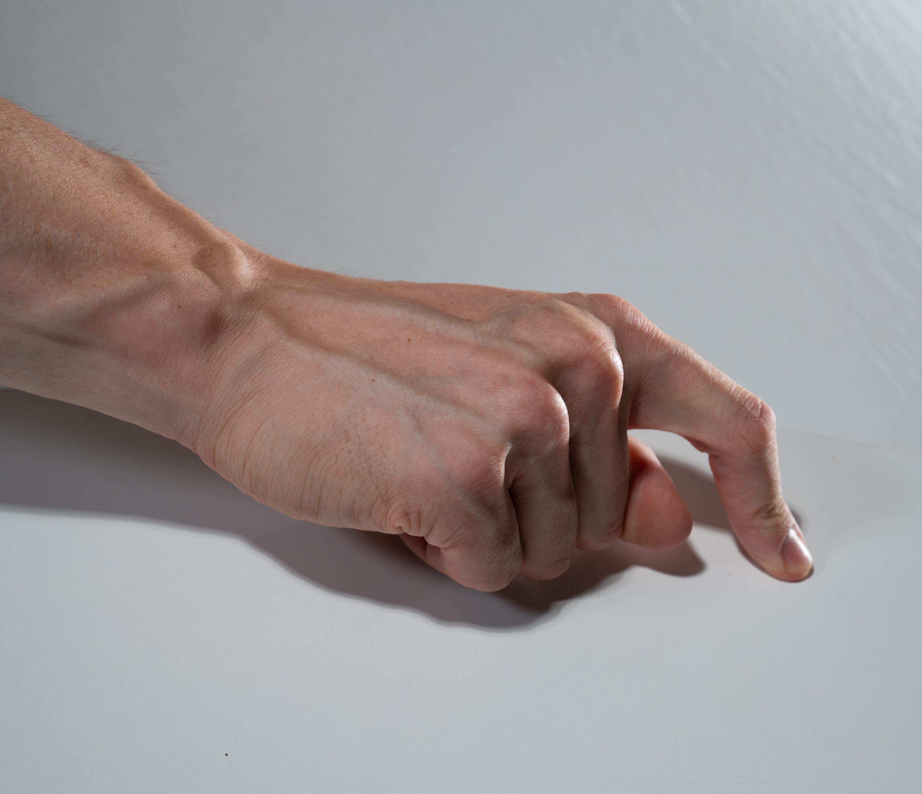 a man's hand is extended on a smooth, light gray surface. the image is taken from above, showing the back of the hand with visible veins. the hand is making a subtle gesture with only the index finger extended while the other fingers are curled in. the lighting is soft and even, creating subtle shadows.