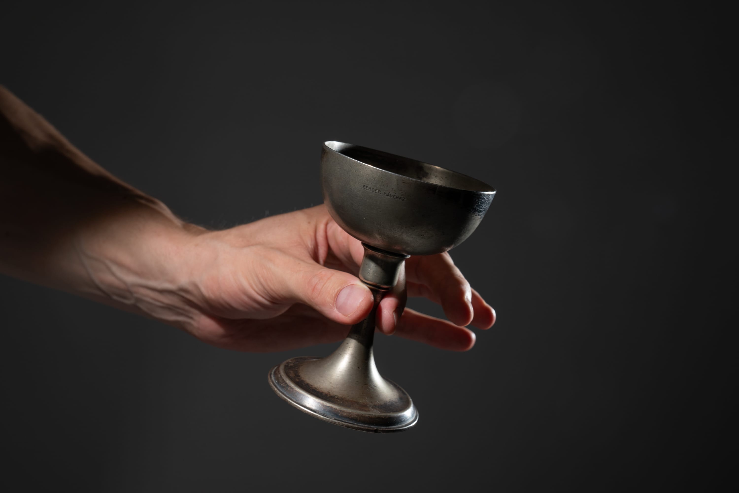 a caucasian male hand is holding a metal cup against a gray background. the cup is old and tarnished. the hand is outstretched as if offering the cup.