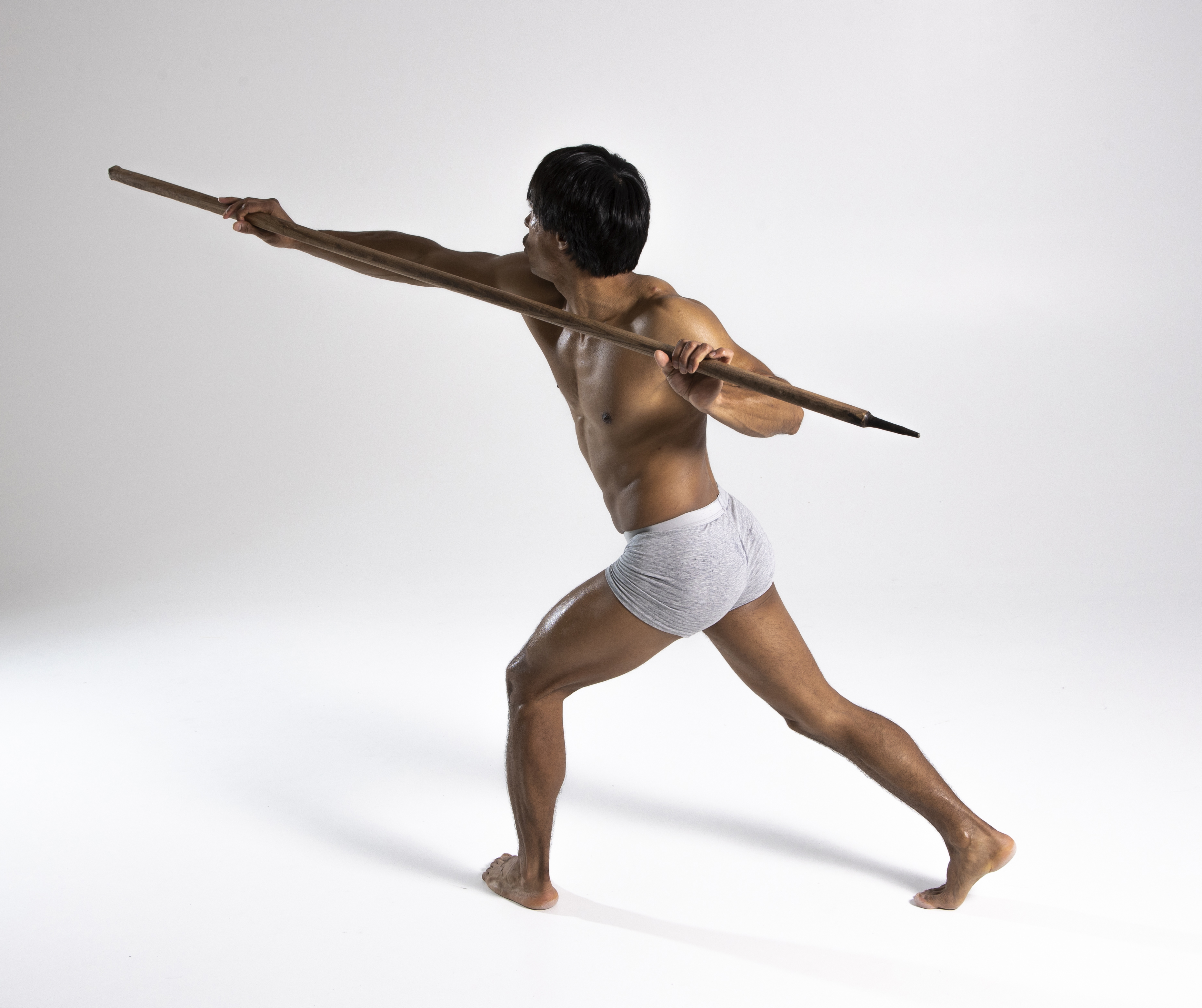 a shirtless, muscular man in a loincloth throws a spear against a white studio background. he is in a dynamic, active pose, suggesting he is a warrior, hunter, or indigenous tribesman.