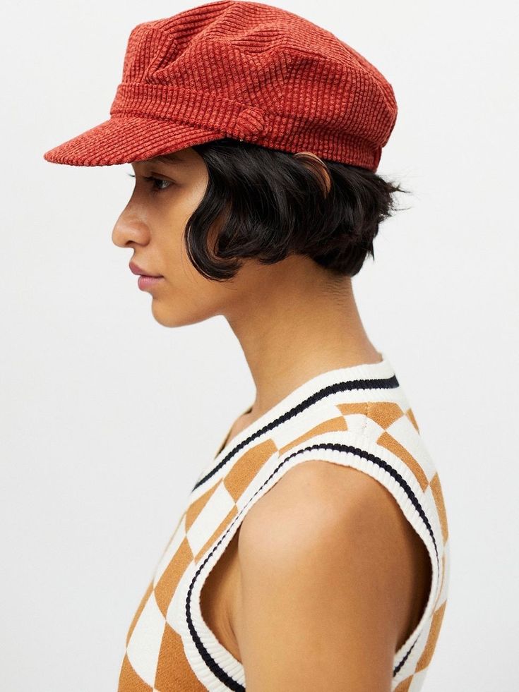 a young woman with short black hair is wearing a red corduroy beret and a white and tan sleeveless sweater. she is facing sideways and looking at the camera. her hair is slightly curled. she is wearing makeup and her lips are slightly parted. she is standing in a studio and the background is white. she has a serious expression on her face.