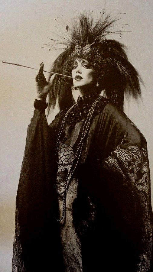 a black and white portrait of a woman in a dramatic and glamorous fashion. she's adorned with a large feathered hat, extravagant jewelry, and a dark flowing gown, creating a mysterious and intriguing image. the pose and expression suggest a sense of elegance and sophistication.