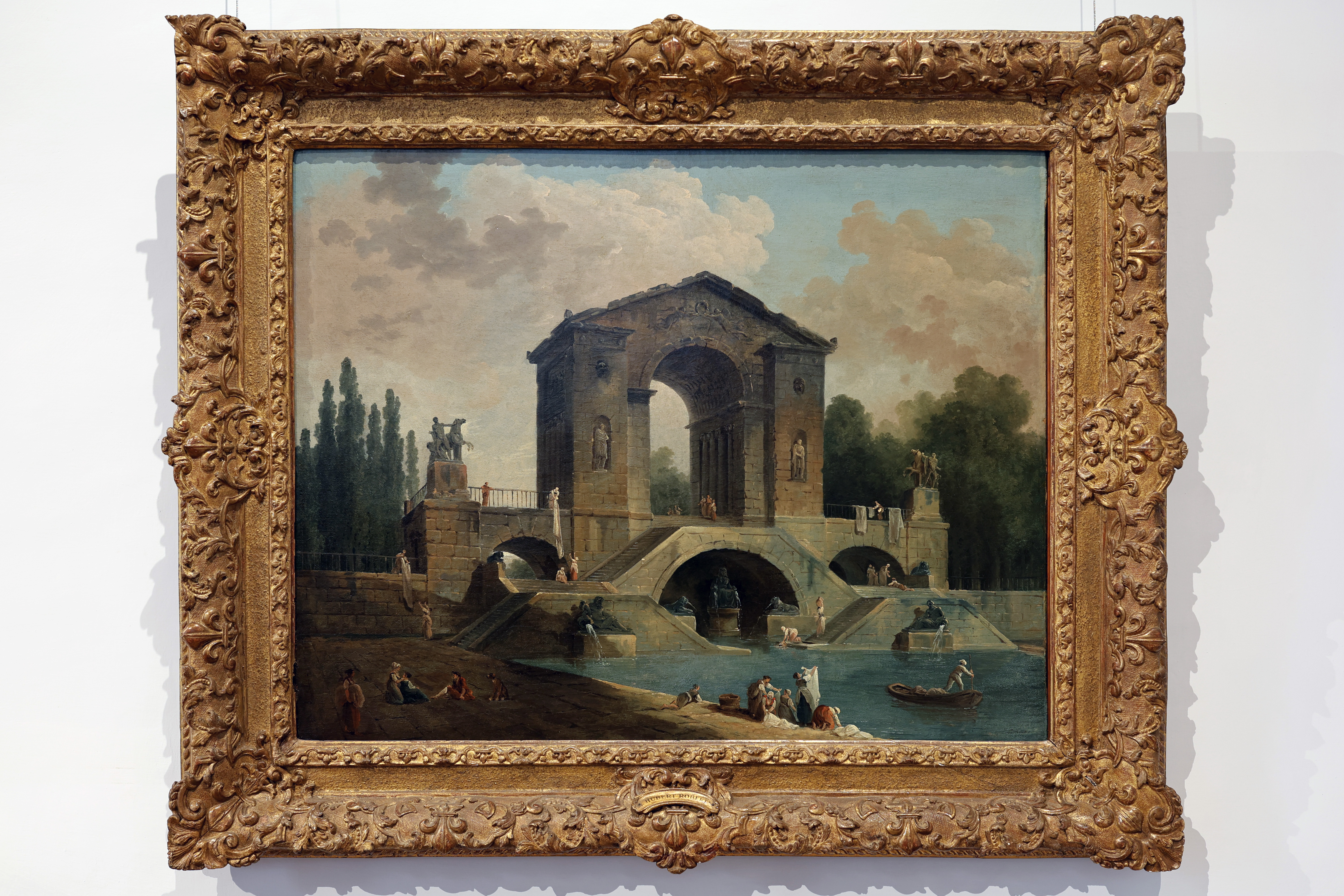 the image is a painting of a landscape scene within an ornate, golden frame. the scene depicts a grand arch structure situated over a bridge and water. various figures are present on the bridge, riverbank, and in boats. the sky is cloudy, and trees populate the background, with statues placed on the bridge.