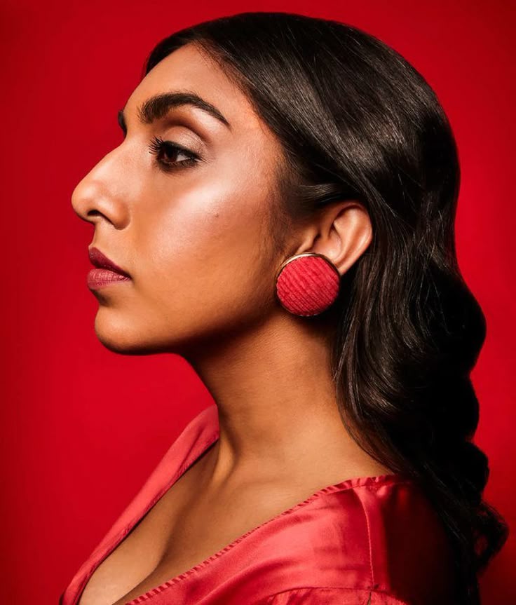 a profile view of a woman with dark hair and dark eyes wearing a red dress and a large red earring against a red background.